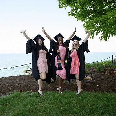 Three Carthage alumna in their caps and gowns in front of Lake Michigan.